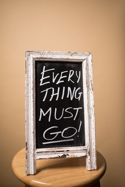 A small, freestanding chalkboard sign with a distressed white wooden frame displaying the handwritten message 'EVERYTHING MUST GO!' in white chalk. The sign is positioned upright on a round wooden tab