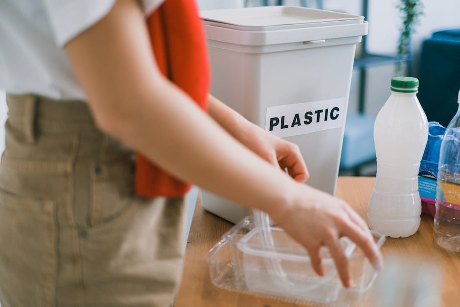A person in beige trousers and a white top, partially visible, is placing a clear plastic container inside a white plastic bin labeled 'PLASTIC' on a light wooden table. The bin is situated in a well-