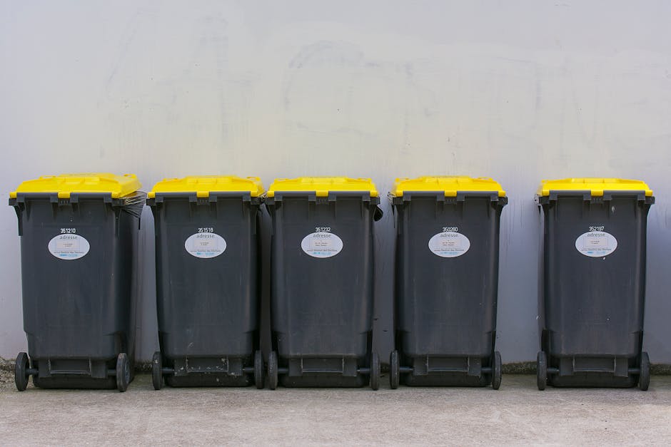 A person in beige trousers and a white top, partially visible, is placing a clear plastic container inside a white plastic bin labeled 'PLASTIC' on a light wooden table. The bin is situated in a well-