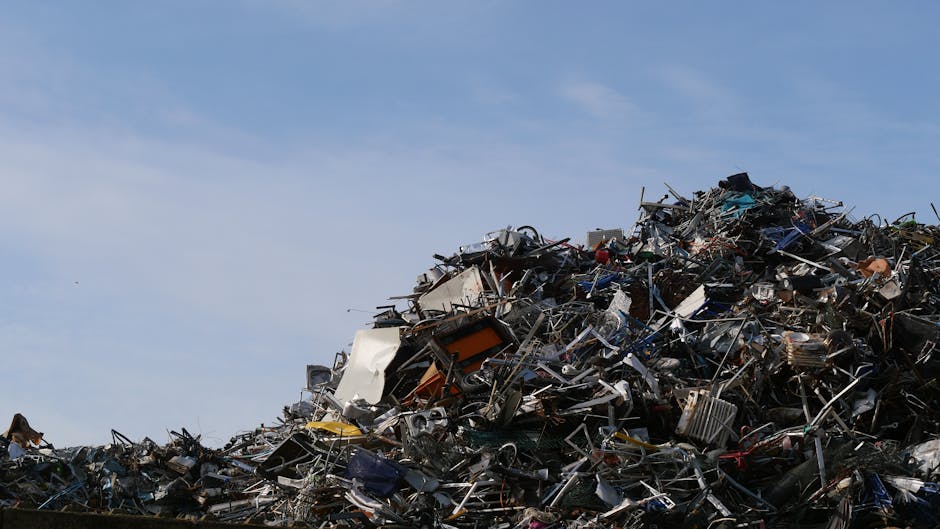 An outdoor scene showing a large pile of assorted discarded household items and rubbish, including broken appliances, plastic containers, and miscellaneous debris. In the foreground, there is a small
